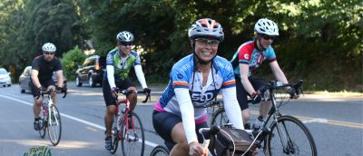 Four bicyclists riding towards and past the camera along a roadside wearing helmets and distance riding gear. The woman on the closest bike is looking at the viewer and grinning.